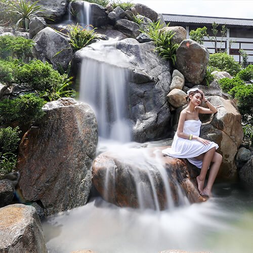 Người phụ nữ ngồi thư giãn bên thác nước nhân tạo giữa khu vườn xanh mát, tạo cảm giác thanh bình và thoải mái.