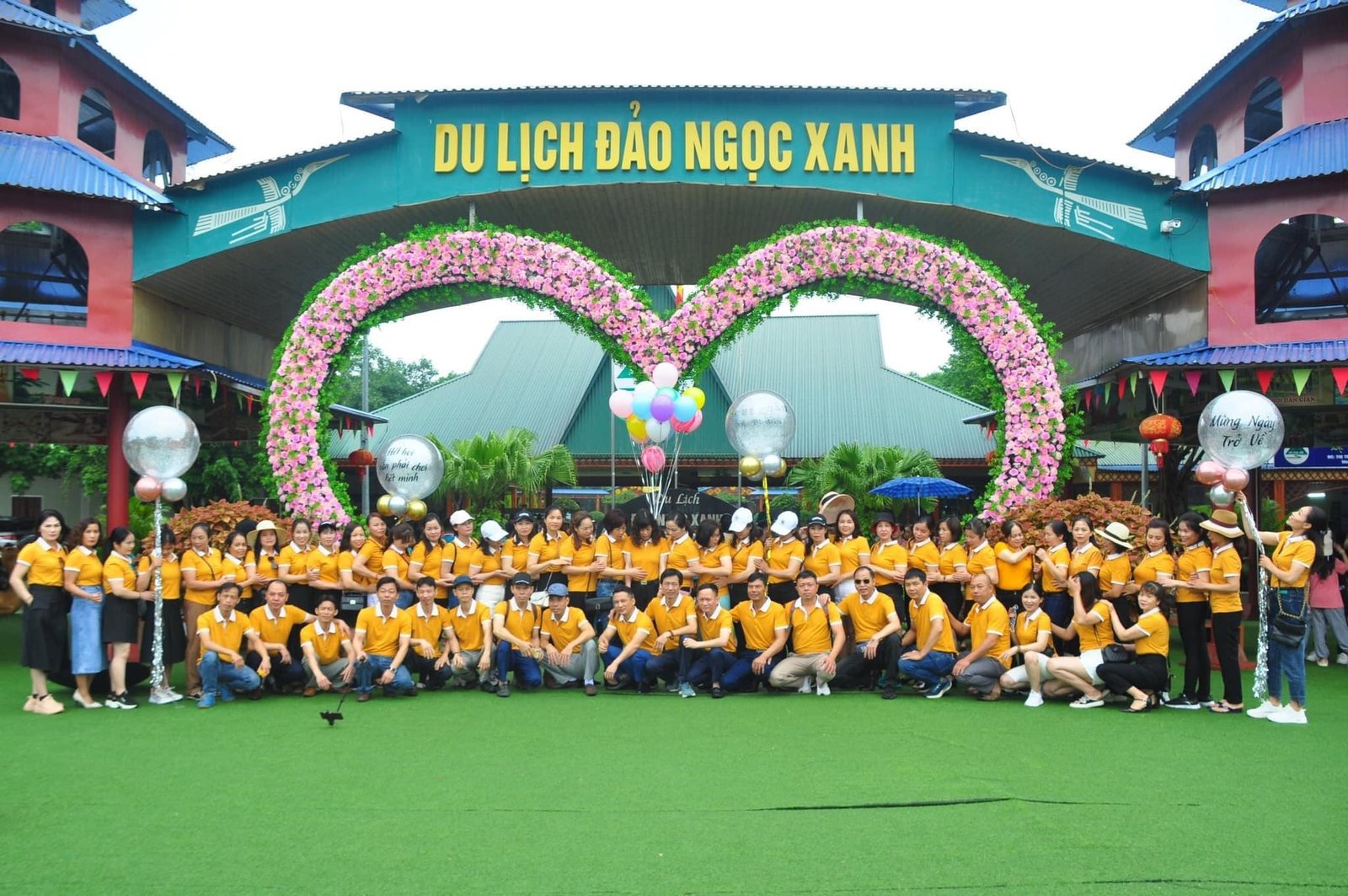 Một nhóm người mặc áo vàng chụp ảnh trước cổng khu du lịch với vòng hoa lớn màu hồng.