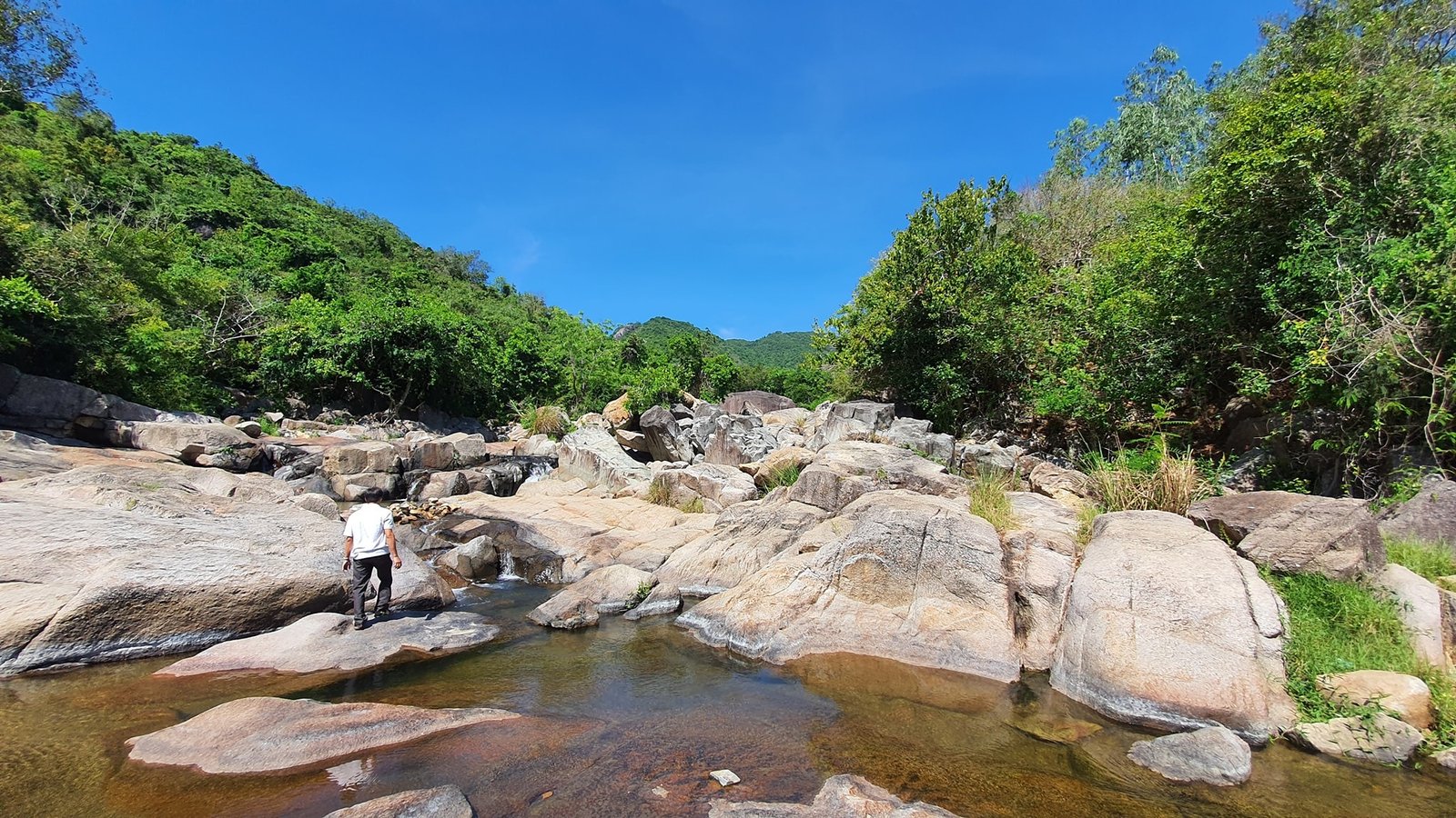 Người đi bộ giữa suối đá trong khu rừng núi xanh tươi dưới bầu trời trong vắt.
