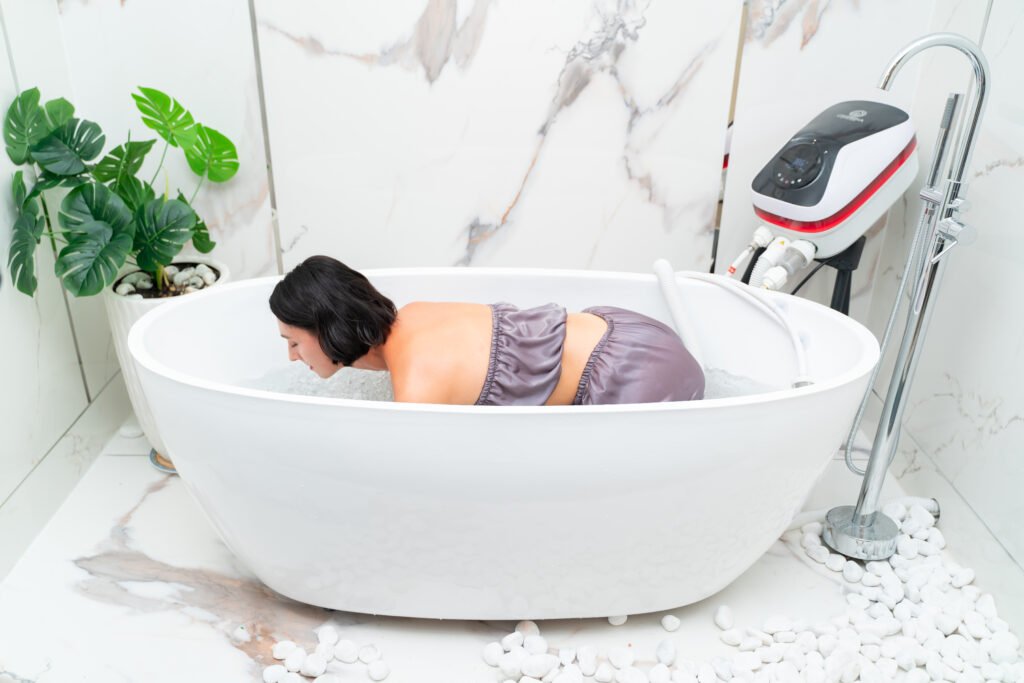 Onsena SPA - Máy Tắm Thủy Liệu Khoáng Nóng Onsen Nhật Bản 94 Người phụ nữ thư giãn trong bồn tắm trắng, bên cạnh là cây xanh, tạo không gian thư giãn hiện đại và thanh lịch.