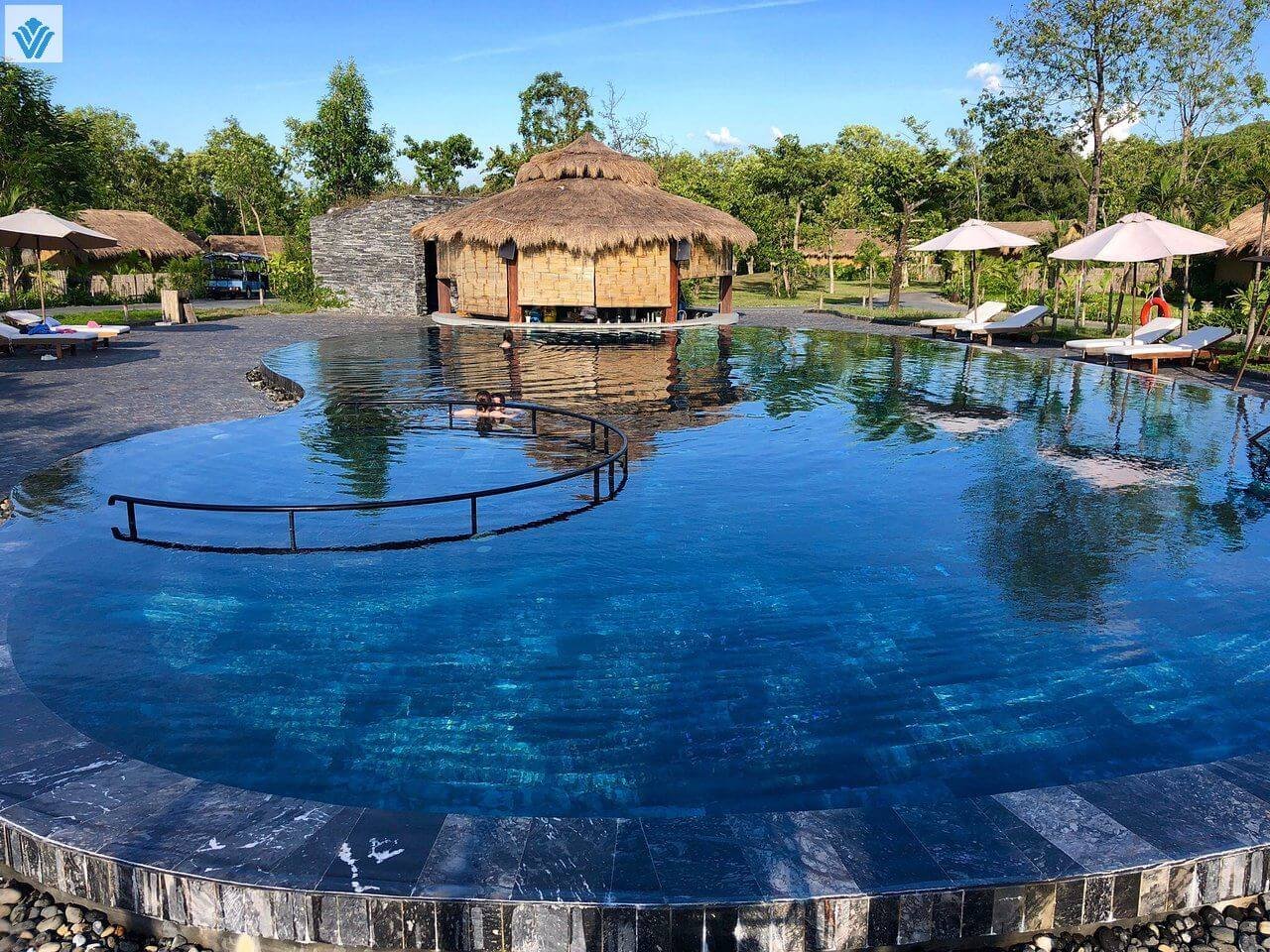 Hồ bơi ngoài trời xanh mát với chòi tranh, ghế nằm thư giãn và cây xanh xung quanh tại resort sang trọng.