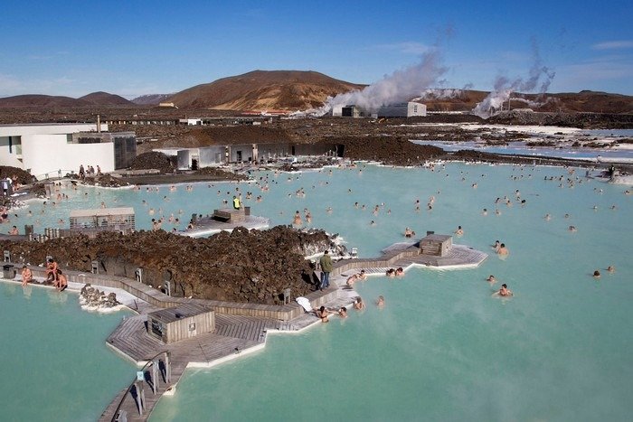 Khám Phá Blue Lagoon - Suối Nước Nóng Tự Nhiên Tuyệt Mỹ Năm 2025 1 Mọi người thư giãn trong một suối nước nóng ngoài trời với khung cảnh núi non và khói bốc lên từ xa.