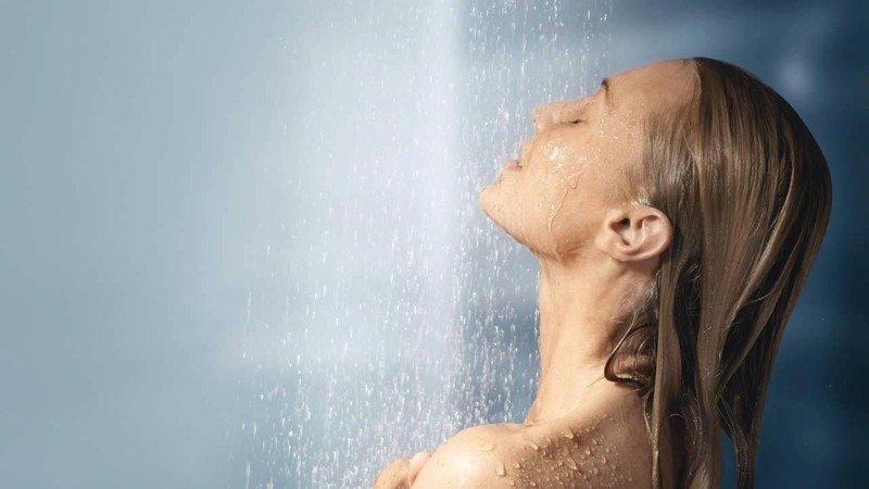 Người phụ nữ thư giãn dưới vòi hoa sen, nước chảy nhẹ nhàng trên tóc và vai, tạo cảm giác sảng khoái và thư thái.