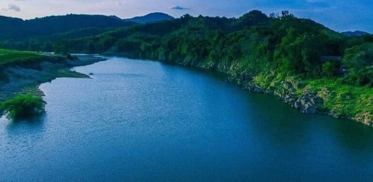 Dòng sông uốn lượn giữa thiên nhiên xanh mát, bao quanh bởi núi đồi và rừng cây, trời trong xanh.