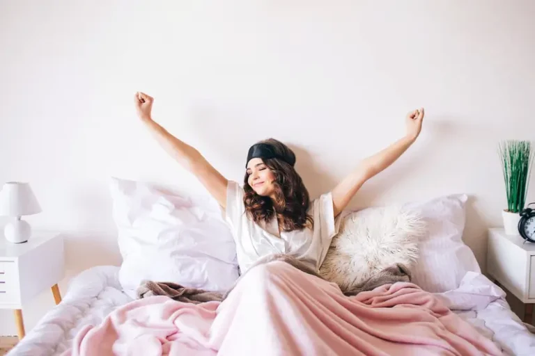 Người phụ nữ vui tươi thức dậy trên giường với chăn hồng và đèn ngủ, cảm giác sảng khoái buổi sáng.