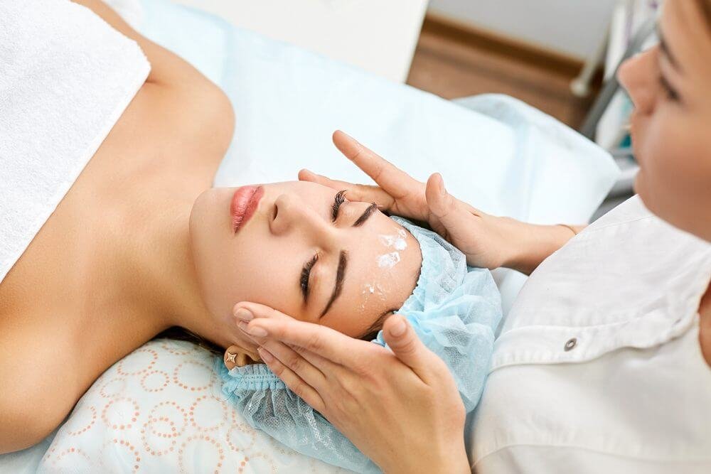 Người phụ nữ thư giãn khi được massage mặt tại spa, đắp khăn và băng đầu màu xanh.