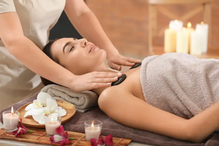 Người phụ nữ được trị liệu đá nóng thư giãn trong spa với nến lung linh và hoa lan trắng.