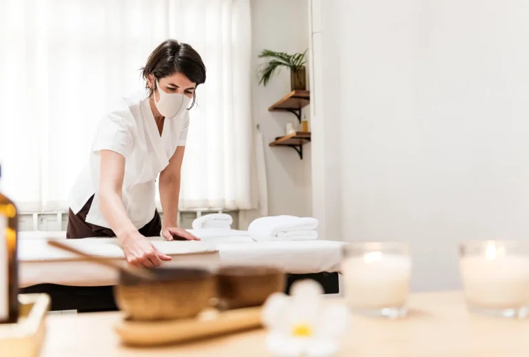 Nhân viên spa đeo khẩu trang chuẩn bị giường massage trong phòng thư giãn với nến và tinh dầu.