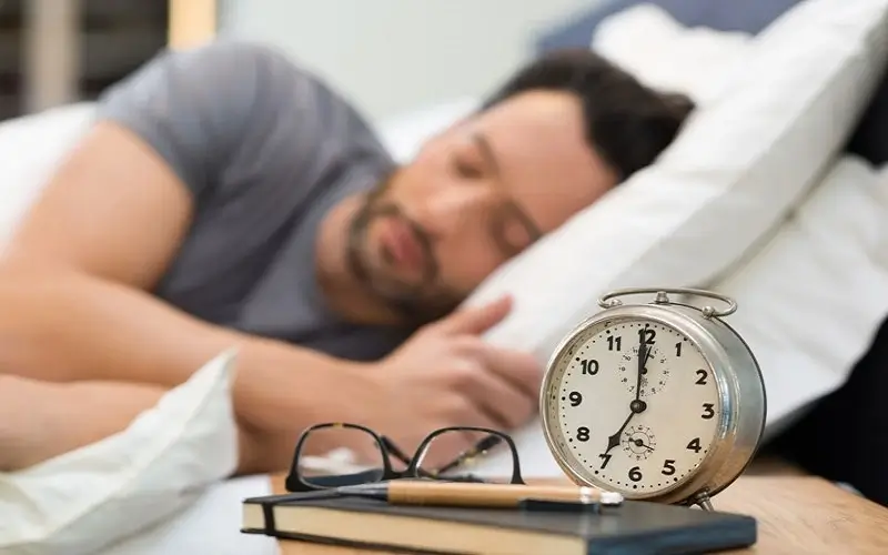 Người đàn ông ngủ say bên cạnh đồng hồ báo thức trên bàn, biểu tượng của giấc ngủ và thời gian nghỉ ngơi.