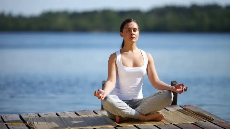 Người phụ nữ tập yoga trong tư thế thiền trên bến tàu, phía trước là hồ nước trong xanh và cây xanh.