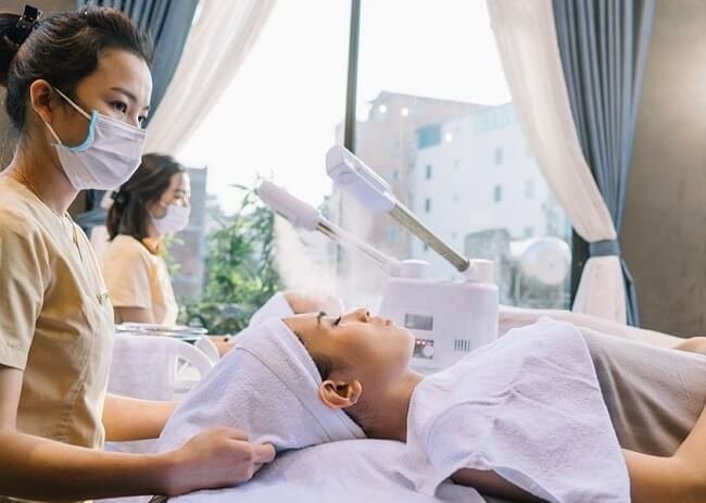 Nhân viên spa đang chăm sóc da mặt cho khách hàng tại thẩm mỹ viện, máy xông hơi đang hoạt động.