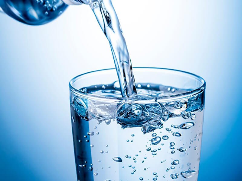 Ly nước trong suốt đang được rót đầy, bong bóng nổi trên bề mặt. Màu xanh dương làm nền tươi mát.