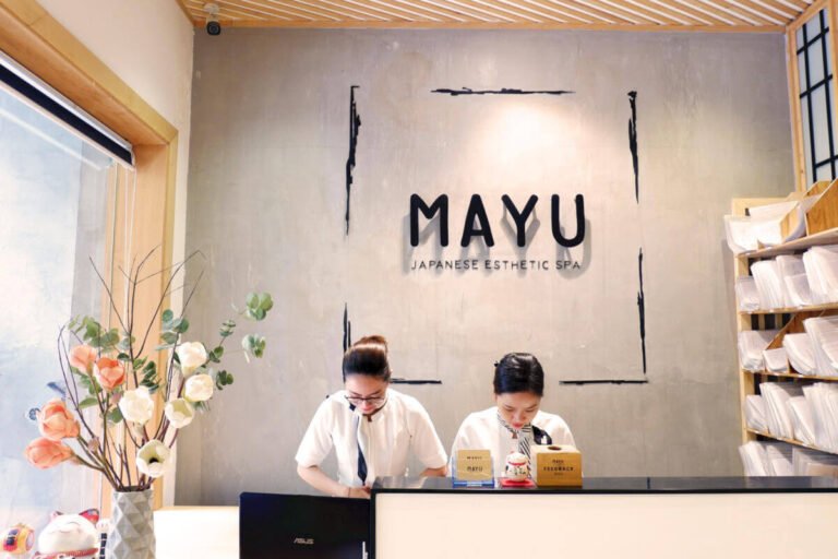 Nhân viên lễ tân tại spa thẩm mỹ Mayu Nhật Bản đang chào khách, với nền hoa và bảng hiệu spa phía sau.