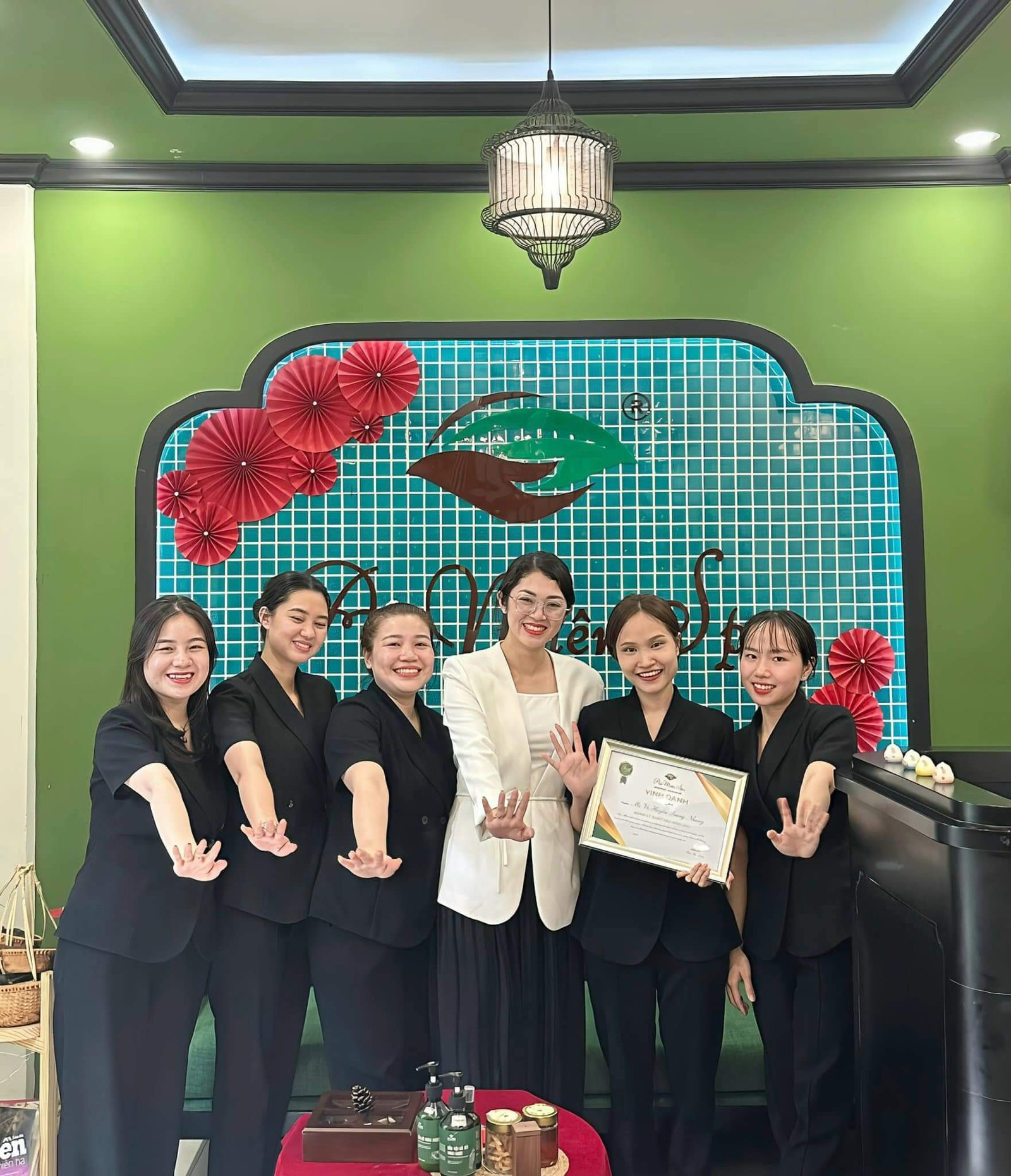 Nhóm phụ nữ cười, mặc đồng phục đen, giơ tay và cầm bằng khen trước logo trên bức tường xanh.