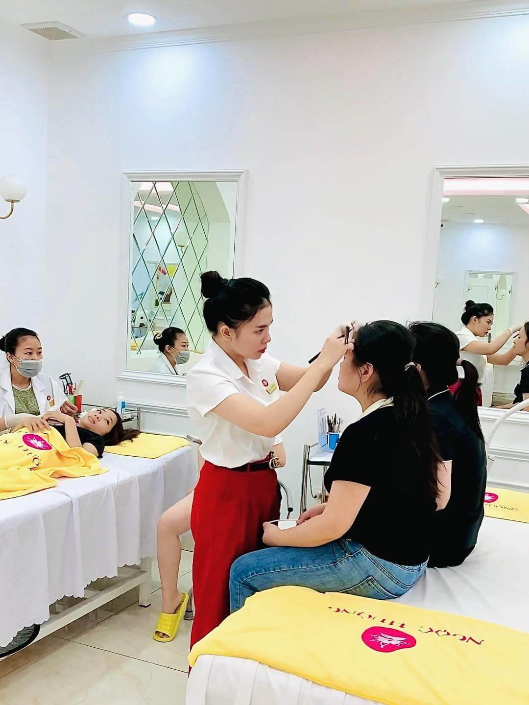 Người phụ nữ đang chăm sóc da mặt cho khách hàng trong spa, với nhiều người khác đang thư giãn xung quanh.