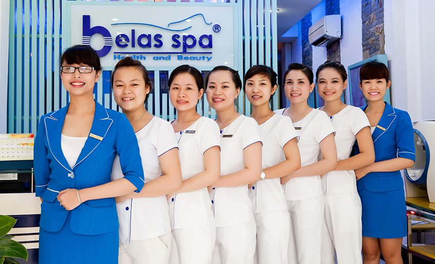 Nhân viên Belas Spa xếp hàng tươi cười, đồng phục trắng xanh, phục vụ sức khỏe và làm đẹp chuyên nghiệp.