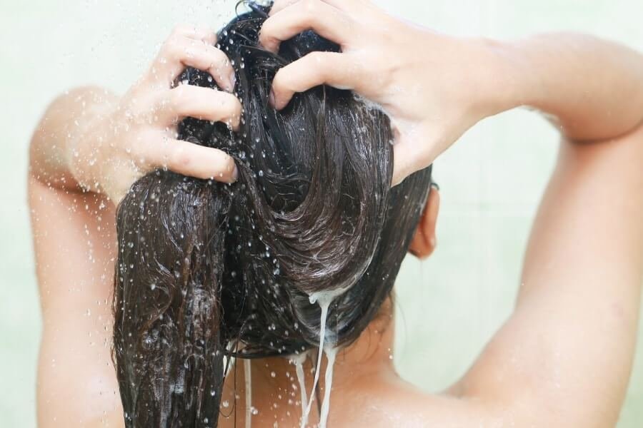 Người phụ nữ đang gội đầu dưới vòi sen, bàn tay thoa dầu gội lên tóc ướt, tạo bọt trắng.