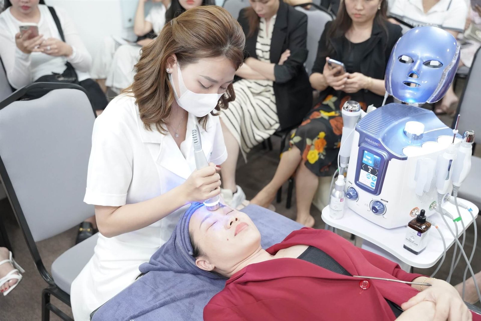 Người phụ nữ đeo khẩu trang đang thực hiện chăm sóc da mặt cho khách hàng trong phòng với thiết bị làm đẹp.
