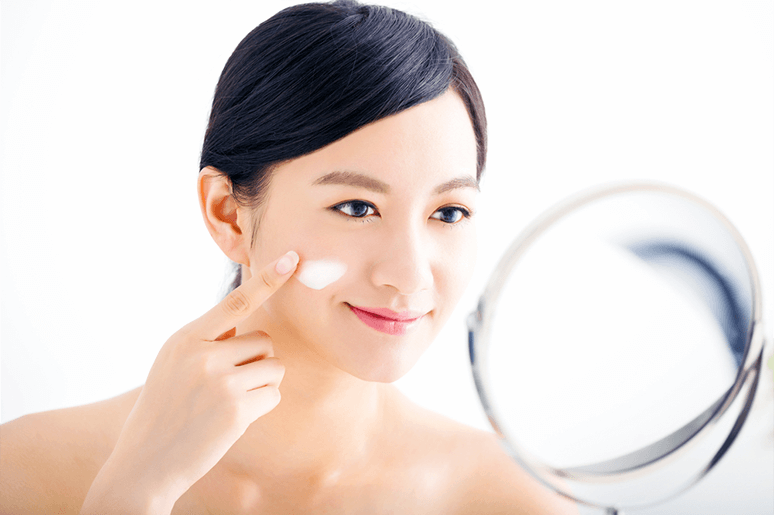 Người phụ nữ thoa kem dưỡng lên má, soi gương mỉm cười trong phòng sáng. Chăm sóc da tự nhiên, sức khỏe làn da.