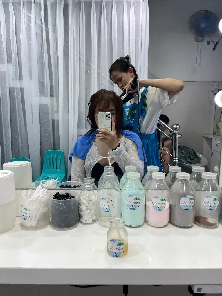 Người phụ nữ chụp ảnh selfie khi đang làm tóc tại salon, trên bàn có nhiều chai đựng sản phẩm chăm sóc tóc.