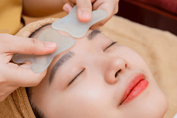 Nhân viên spa thực hiện trị liệu gua sha trên trán khách hàng, giúp thư giãn và cải thiện tuần hoàn máu.