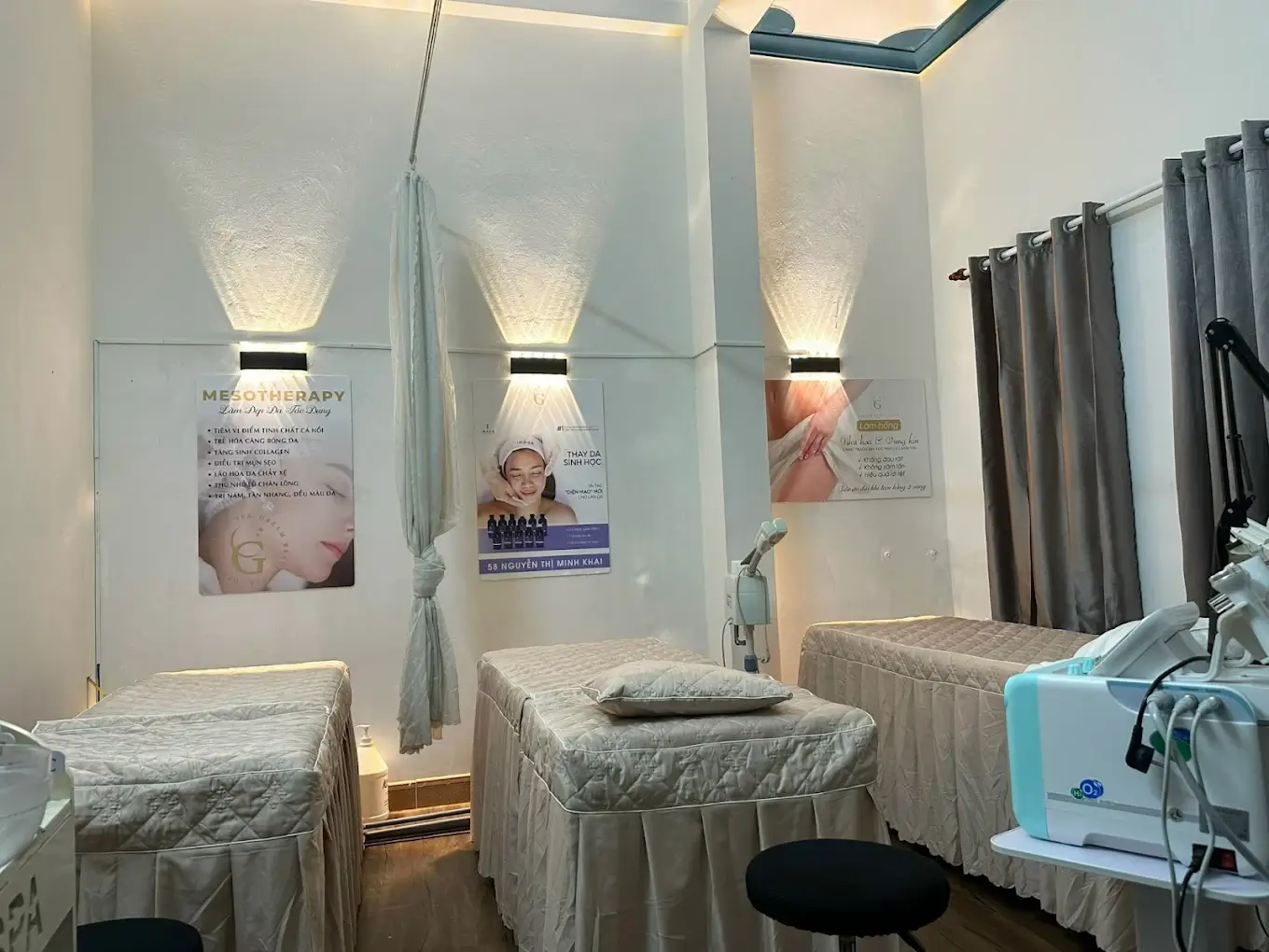 Phòng trị liệu thẩm mỹ với giường massage, máy móc chuyên dụng và áp phích liệu trình, không gian sáng sủa.