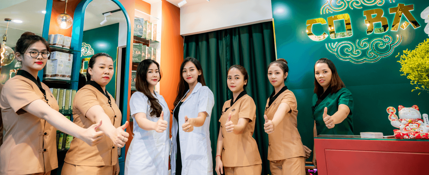 Nhóm nhân viên Cô Ba Massage tạo dáng giơ ngón tay cái tại quầy tiếp tân, mặc đồng phục chuyên nghiệp.