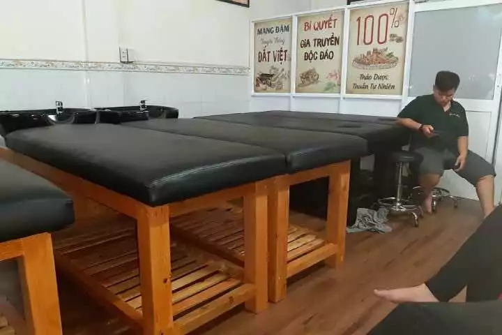 Phòng mát xa với giường gỗ, bảng hiệu thảo dược, người ngồi dùng điện thoại, không gian thư giãn và thoải mái.