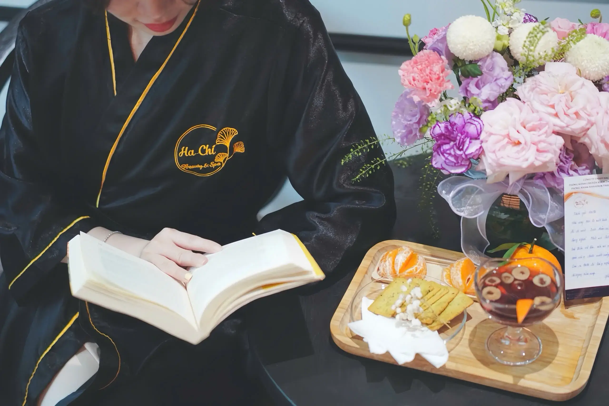 Người phụ nữ đọc sách trong áo choàng spa, bên cạnh là đĩa trái cây và hoa tươi.