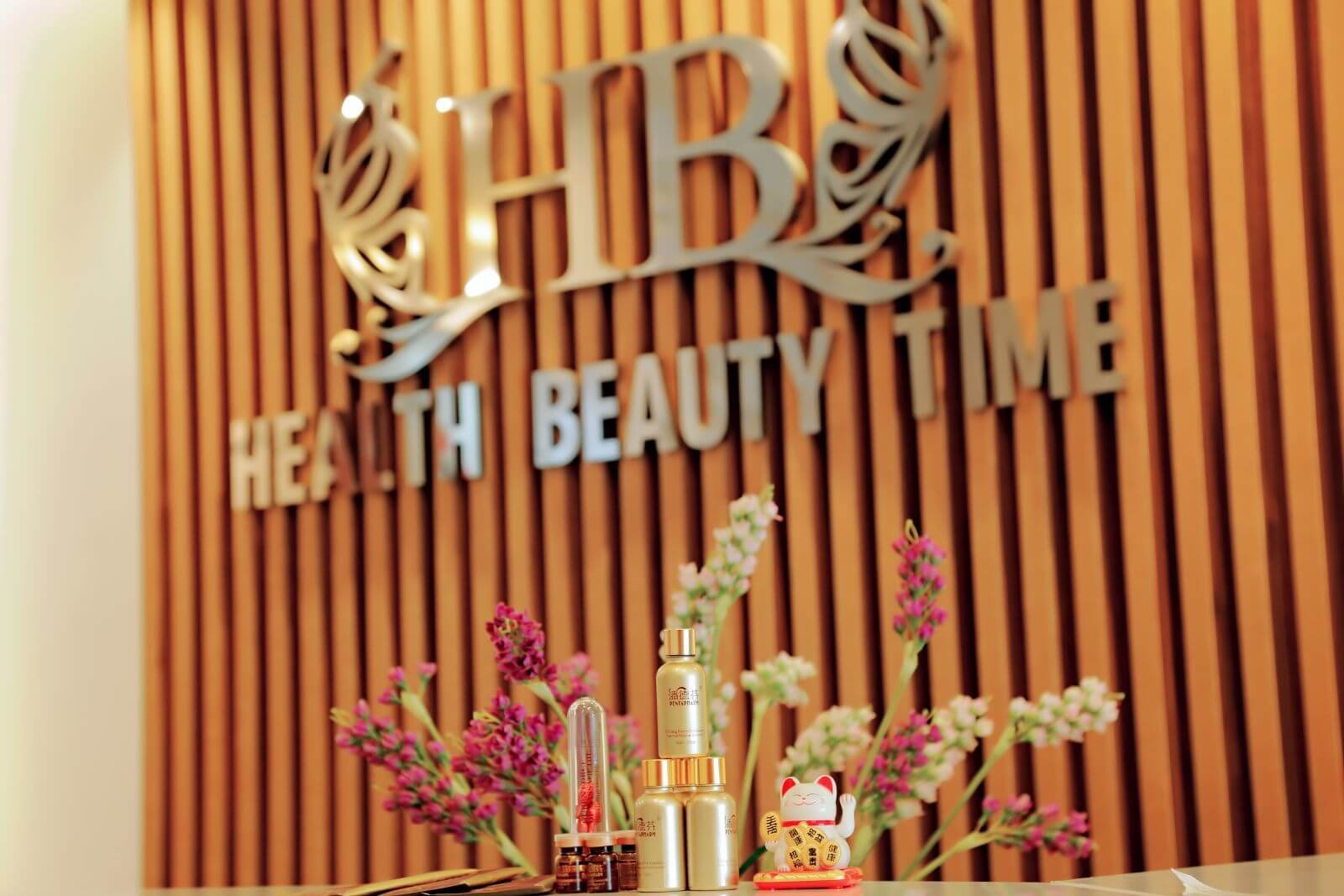 Sản phẩm mỹ phẩm trang trí trước mặt bảng hiệu Health Beauty Time với hoa và tượng mèo may mắn.