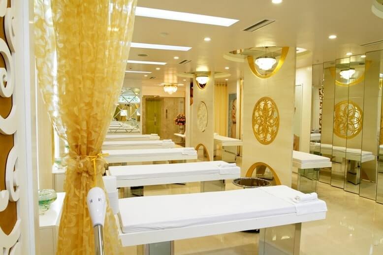 Phòng spa sang trọng với giường massage, rèm vàng, ánh sáng dịu và gương trang trí trên tường.