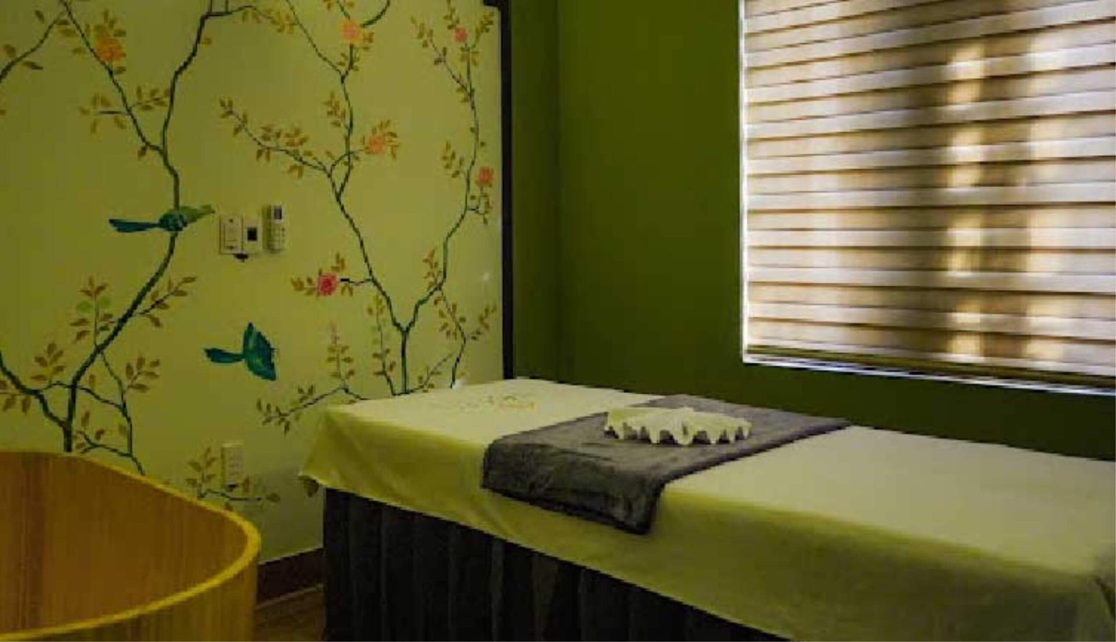 Phòng mát-xa với giường có ga trải trắng, bức tường trang trí hoa lá, và cửa sổ với rèm kéo.