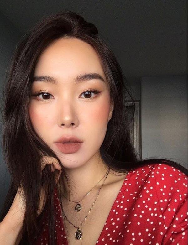 Người phụ nữ có tóc đen và làn da trắng đang chạm tay vào mặt, mặc áo đỏ chấm bi, ánh sáng nhẹ nhàng.