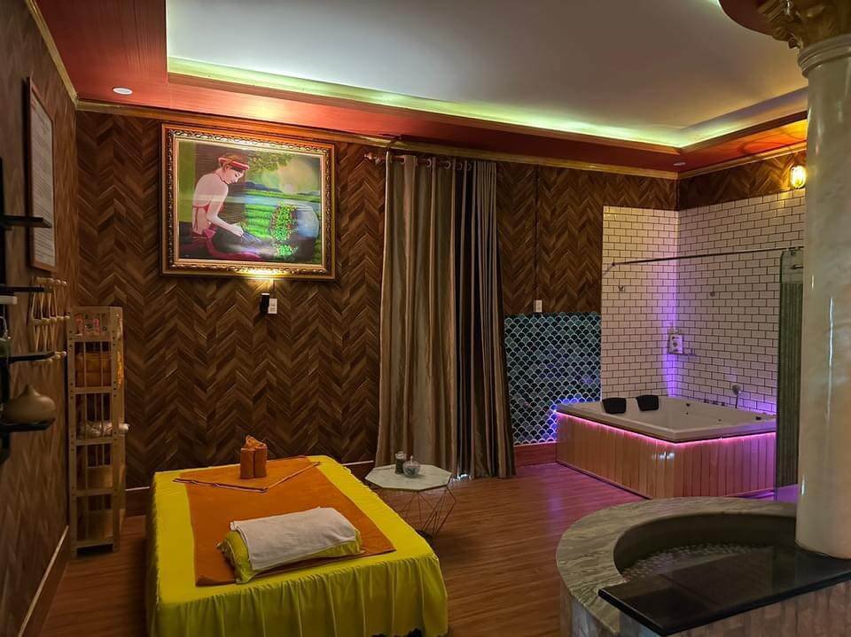 Phòng spa với giường massage, bồn tắm và tranh treo tường. Nội thất ốp gỗ và gạch, ánh sáng dịu nhẹ.