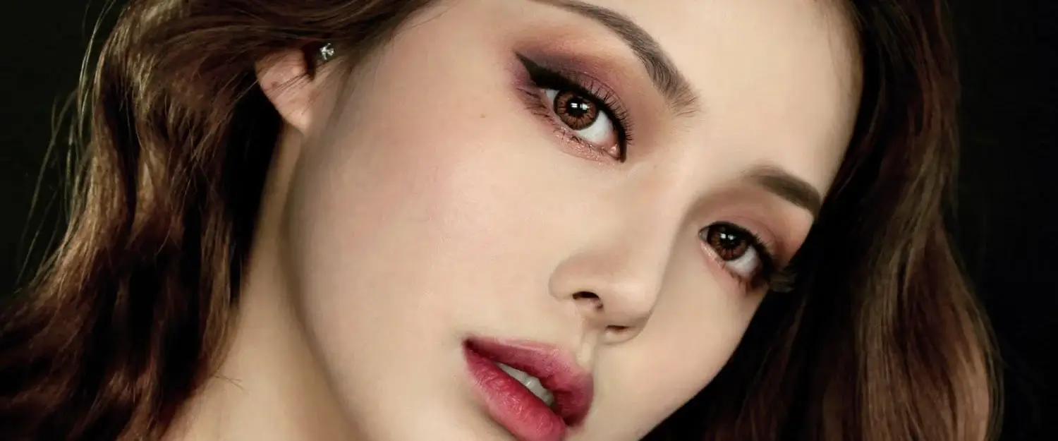 Người phụ nữ với lối trang điểm mắt sắc sảo và đôi môi đỏ hồng đẹp nổi bật, mái tóc nâu gợn sóng nhẹ.