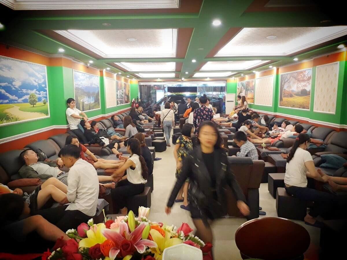 Phòng massage chân đông đúc, nhiều khách thư giãn, nhân viên phục vụ tận tâm trong không gian ấm cúng.