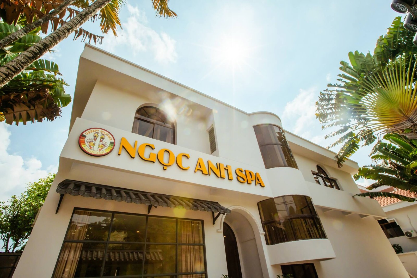 Tòa nhà spa màu trắng với tên trên biển hiệu, bao quanh bởi cây xanh, trời nắng trong xanh.
