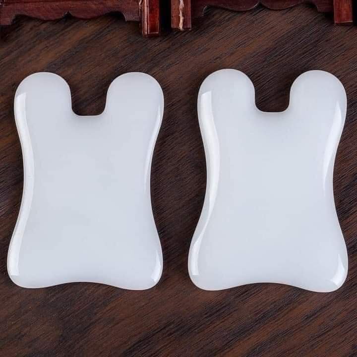 Hai dụng cụ gua sha màu trắng trên nền gỗ, dùng để massage và thư giãn cơ thể.