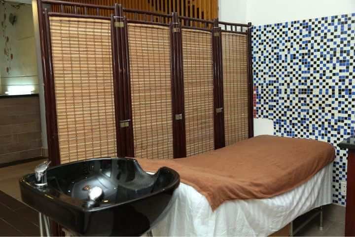 Giường massage với chăn nâu, bên cạnh là bồn gội đầu và vách ngăn tre, tường có gạch lát hoa văn.