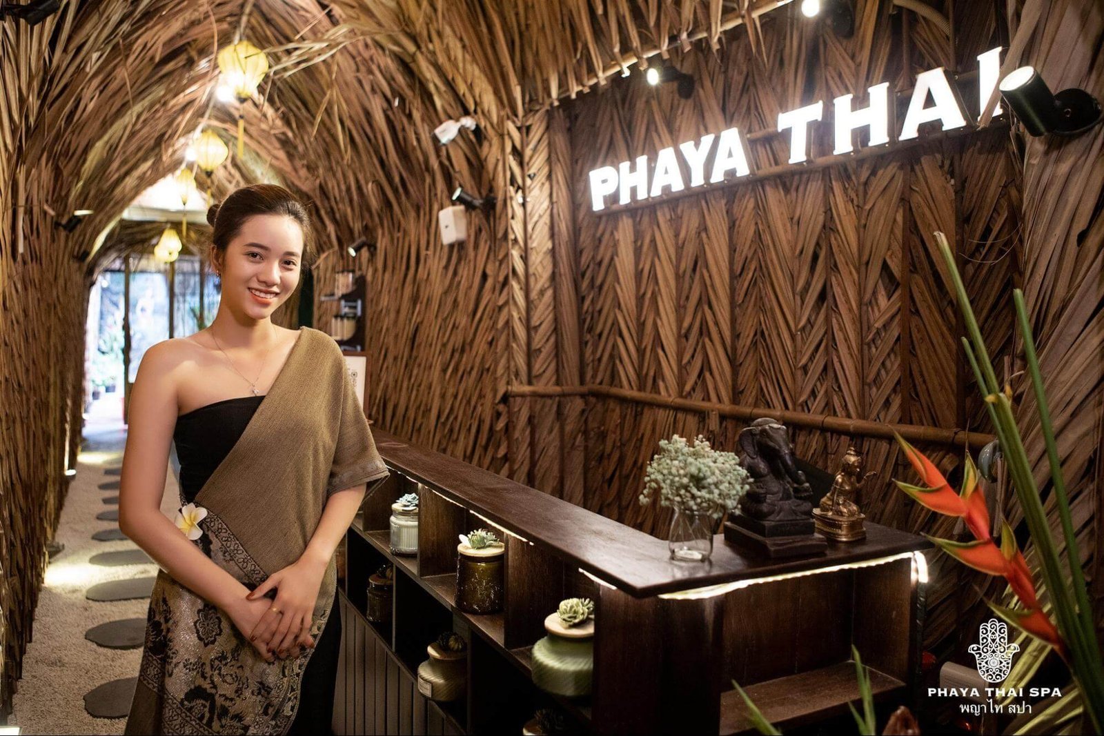 Nhân viên tại spa Phaya Thai, mặc trang phục truyền thống, đứng trước bàn lễ tân với nền trang trí bằng lá cọ.