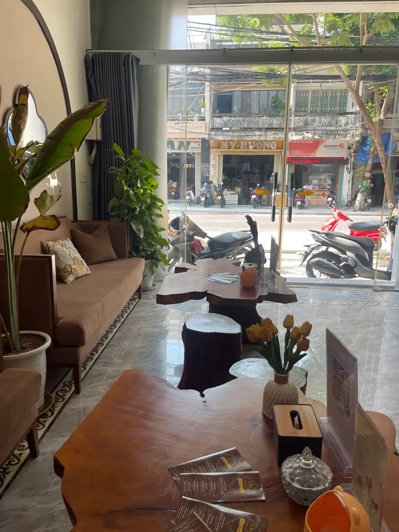 Quán cà phê nội thất gỗ sang trọng, cửa kính lớn, cây xanh, xe máy ngoài đường.