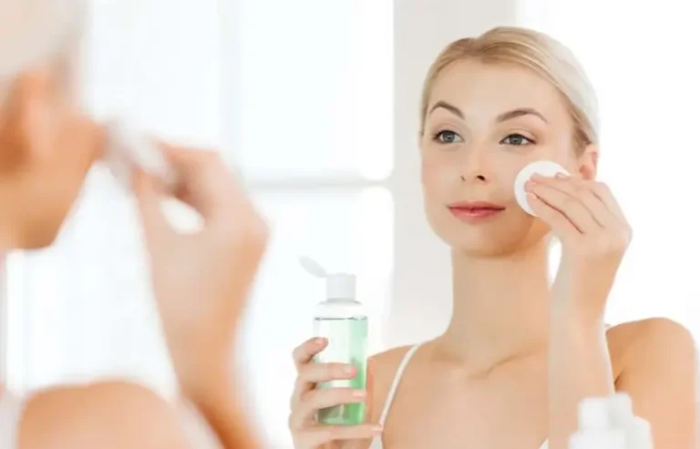 Người phụ nữ chăm sóc da mặt với bông tẩy trang và chai nước hoa hồng trước gương.
