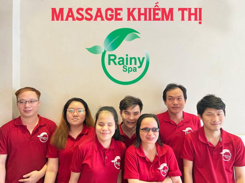 Nhóm nhân viên Massage Khiếm Thị Rainy Spa mặc áo đỏ, đứng bên dưới logo xanh.