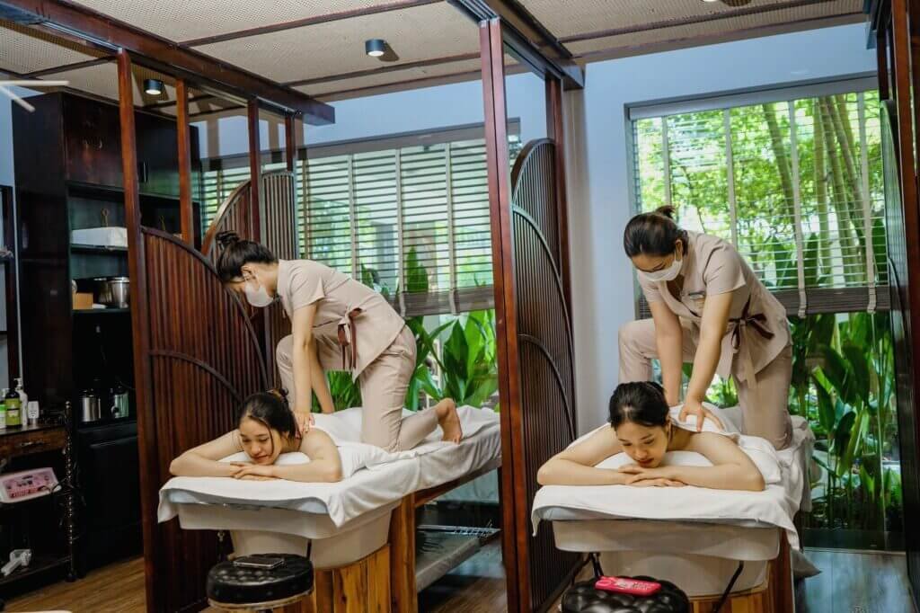 Hai khách hàng đang tận hưởng liệu pháp massage thư giãn tại spa với ánh sáng tự nhiên và cây xanh xung quanh.