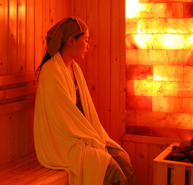 Người ngồi trong phòng xông hơi gỗ, quấn khăn và áo choàng, ánh sáng vàng từ tường muối tạo không gian thư giãn.