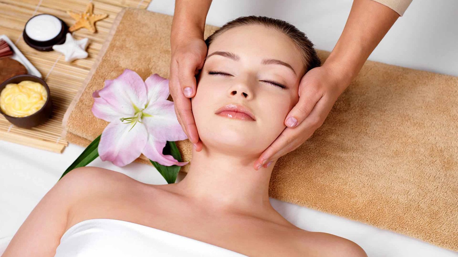 Người đang được mát-xa mặt với hoa và dụng cụ chăm sóc da xung quanh trên khăn mềm màu nâu.