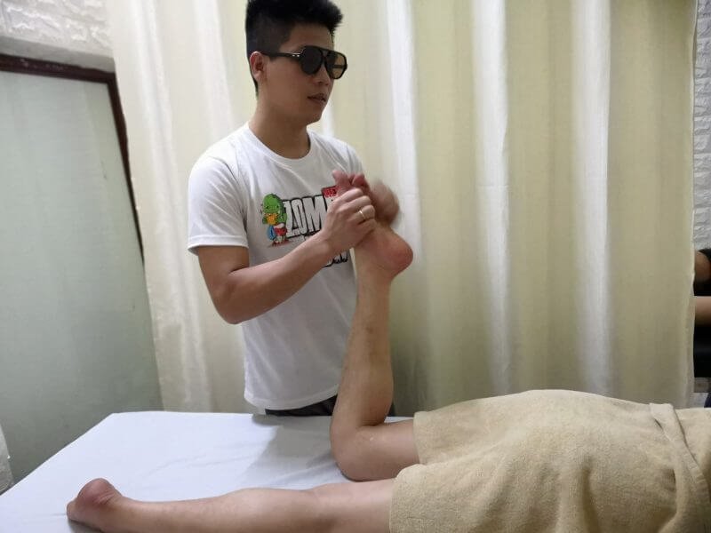Người đàn ông đeo kính mát đang mát-xa chân cho một người khách nằm trên giường, dùng khăn che phần thân.