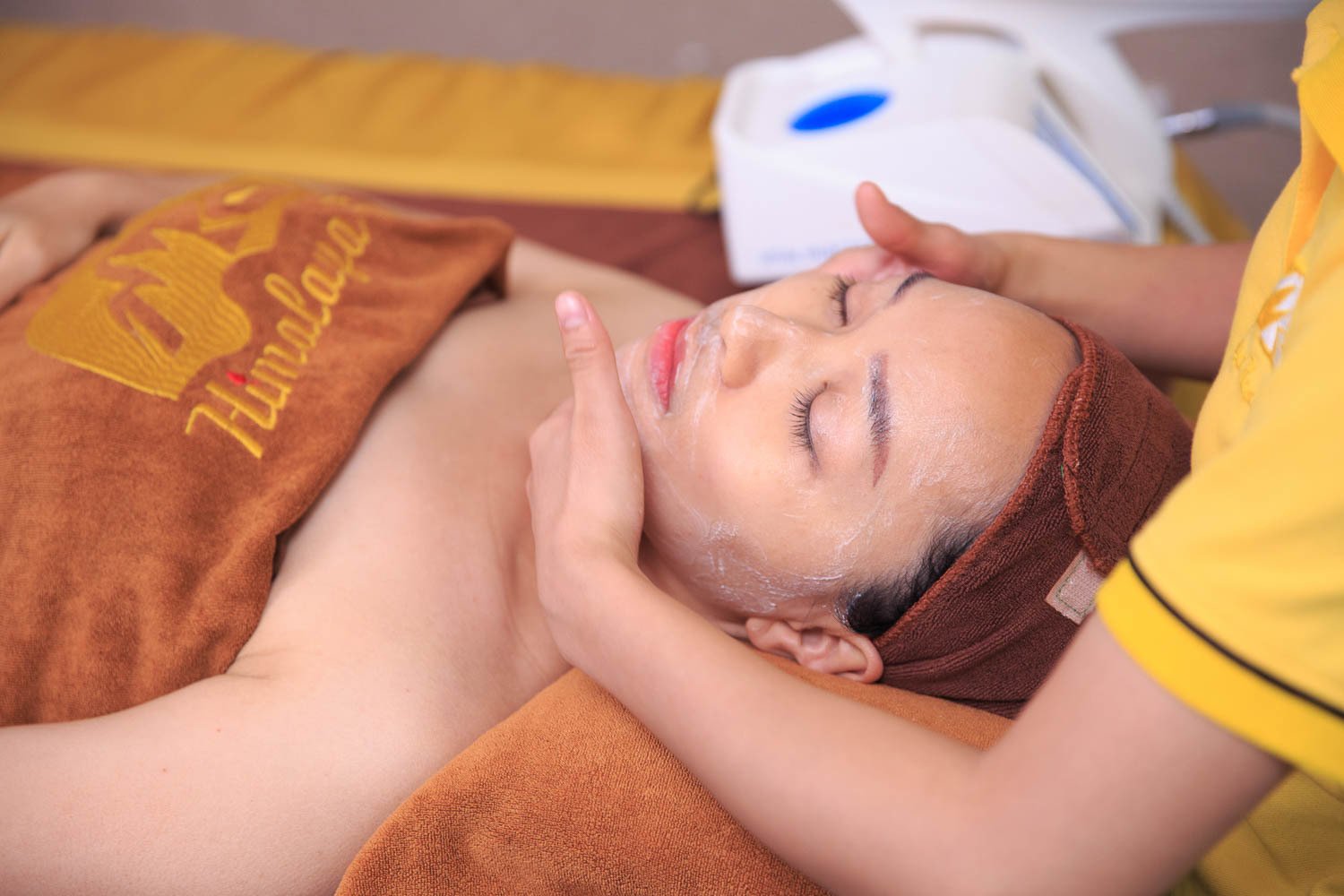 Người đang được mát-xa mặt tại spa, quấn khăn nâu, có sản phẩm dưỡng da trên mặt.
