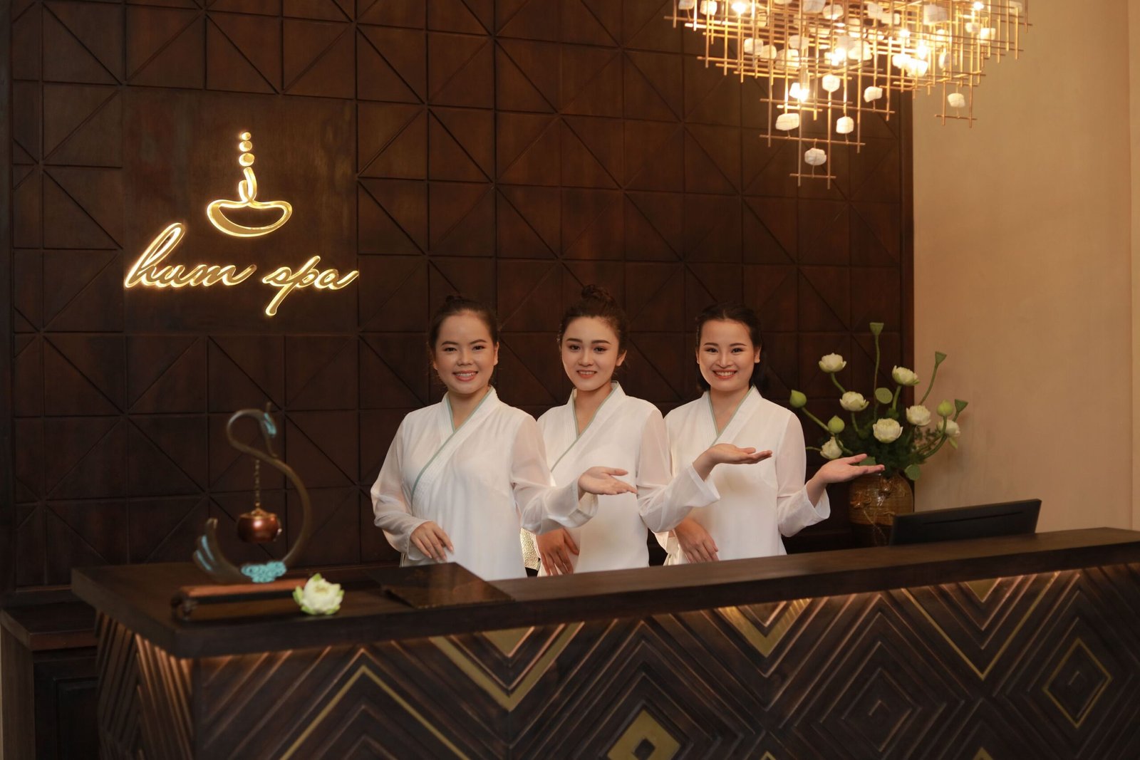 Ba nhân viên spa trong trang phục trắng chào đón khách tại lễ tân, bên cạnh đèn chùm sang trọng.