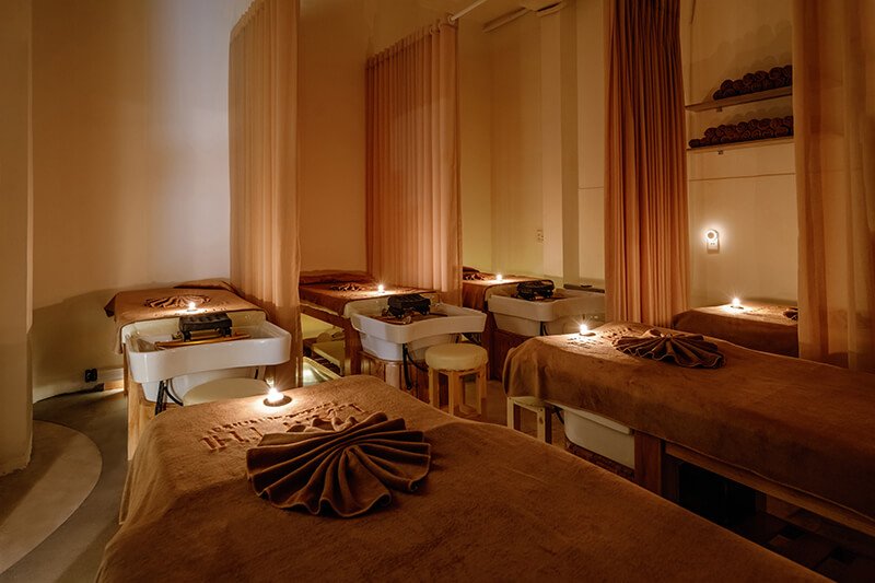 Phòng spa ấm cúng với giường massage, ánh nến dịu nhẹ, rèm và nội thất màu nâu thư giãn.